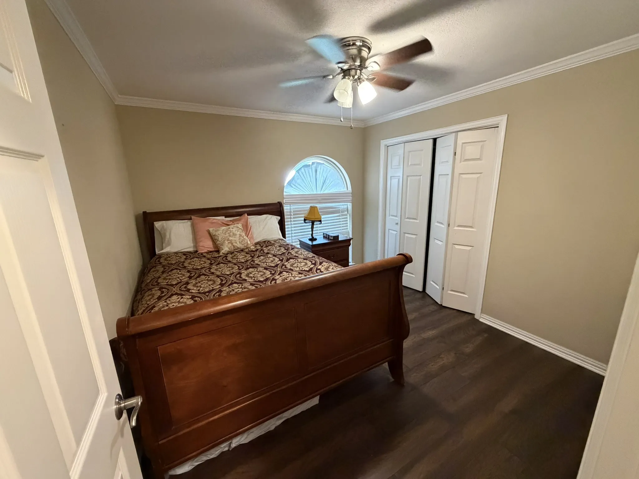 Crown molding, dark wood-finish flooring, ceiling fan with integrated lighting, arched window with privacy blinds, and bifold closet doors