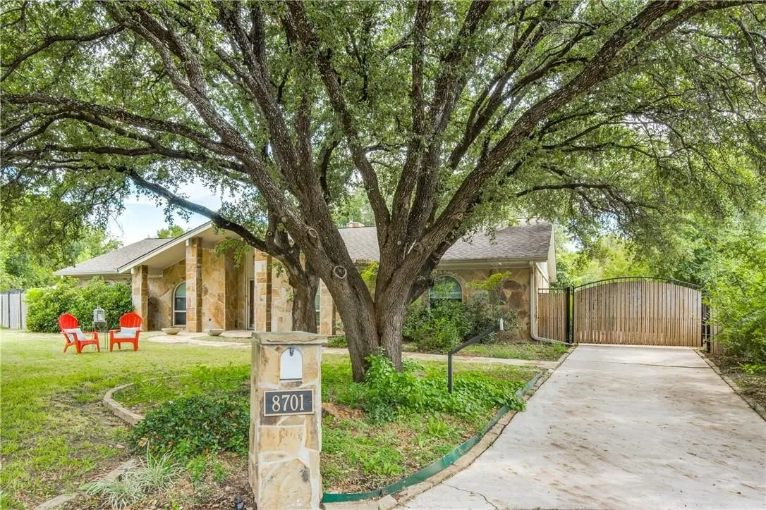 Stone facade, mature trees, gated driveway, stone mailbox with visible address, covered front entry