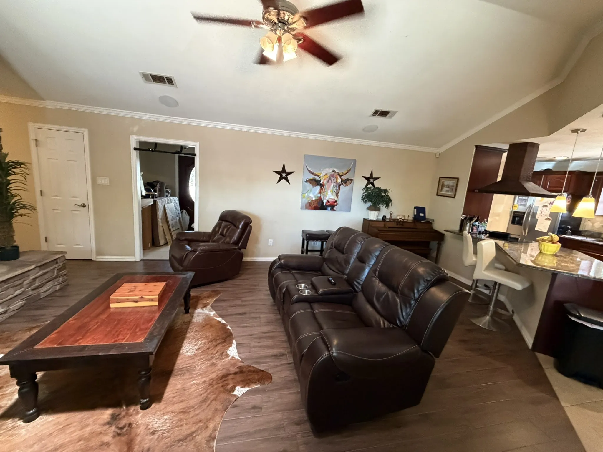 Wood-finish flooring, ceiling fan with integrated lighting, recessed ceiling speakers, crown molding, and a kitchen island with pendant lighting
