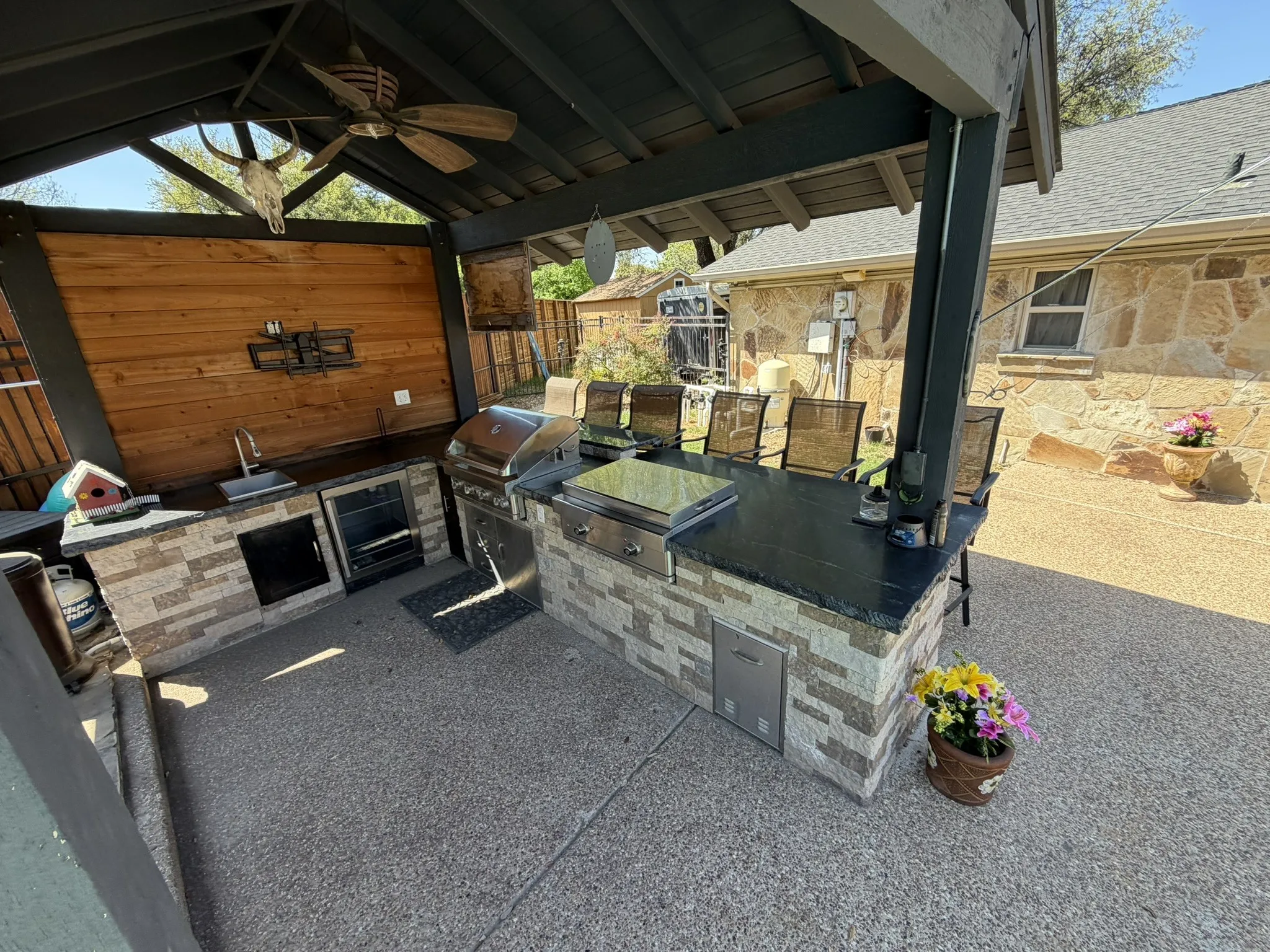 Covered outdoor kitchen featuring a built-in grill, integrated sink, and stone-clad base with countertop seating