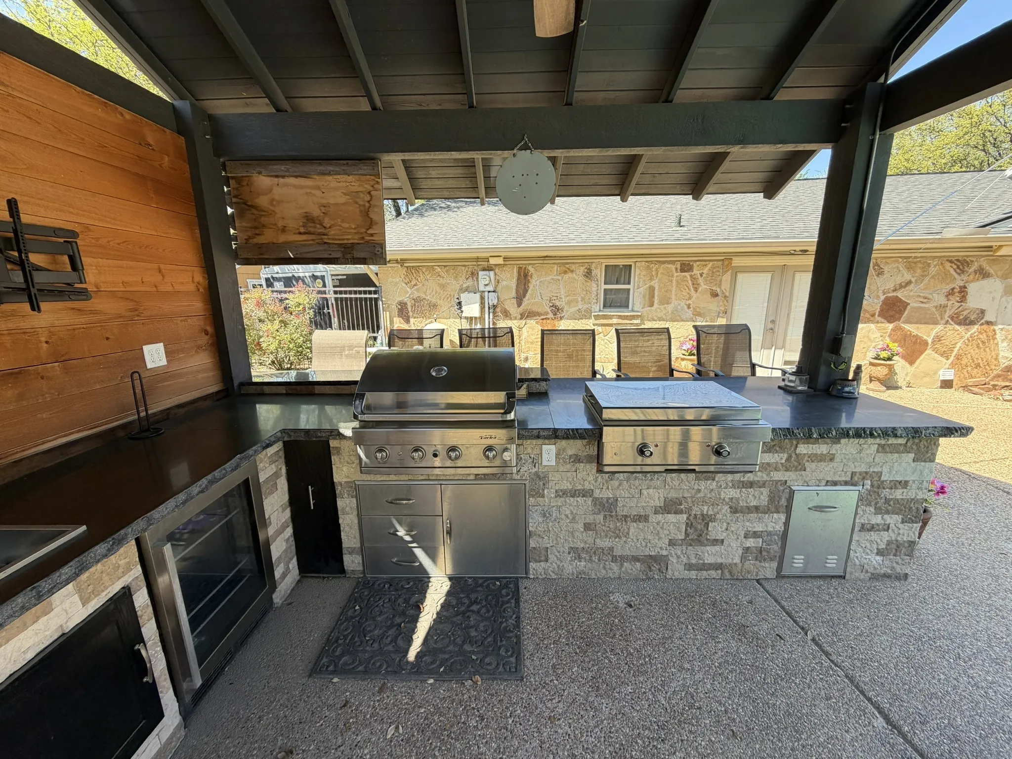 Outdoor kitchen featuring a built-in grill, stone-clad island, dark countertops, and a wood-paneled accent wall