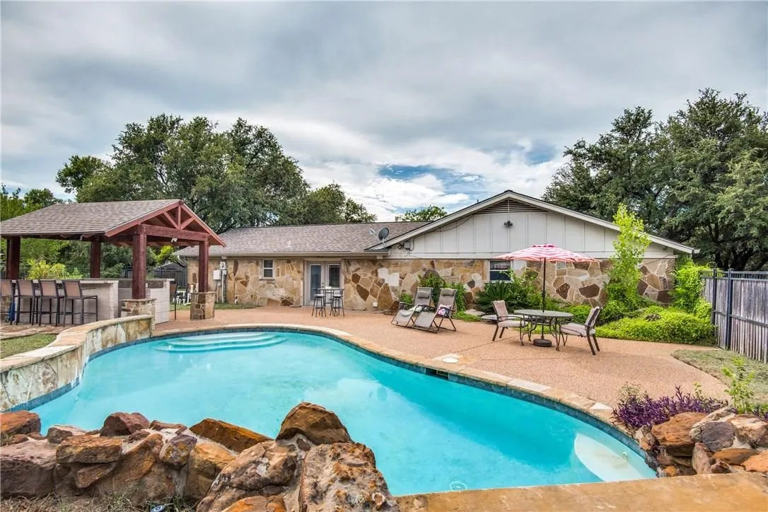 Freeform swimming pool with rock accents and integrated steps, complemented by an outdoor bar under a wooden pergola, a stone-clad exterior, and a paved patio area