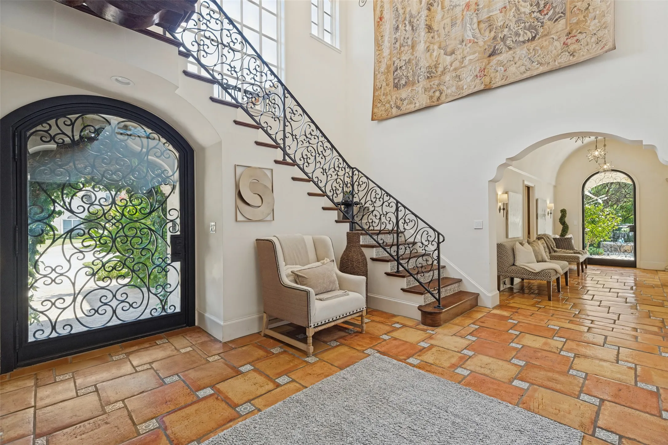 Arched entryway featuring an ornate wrought iron and glass door, terracotta tile flooring, and a grand staircase with wrought iron railing