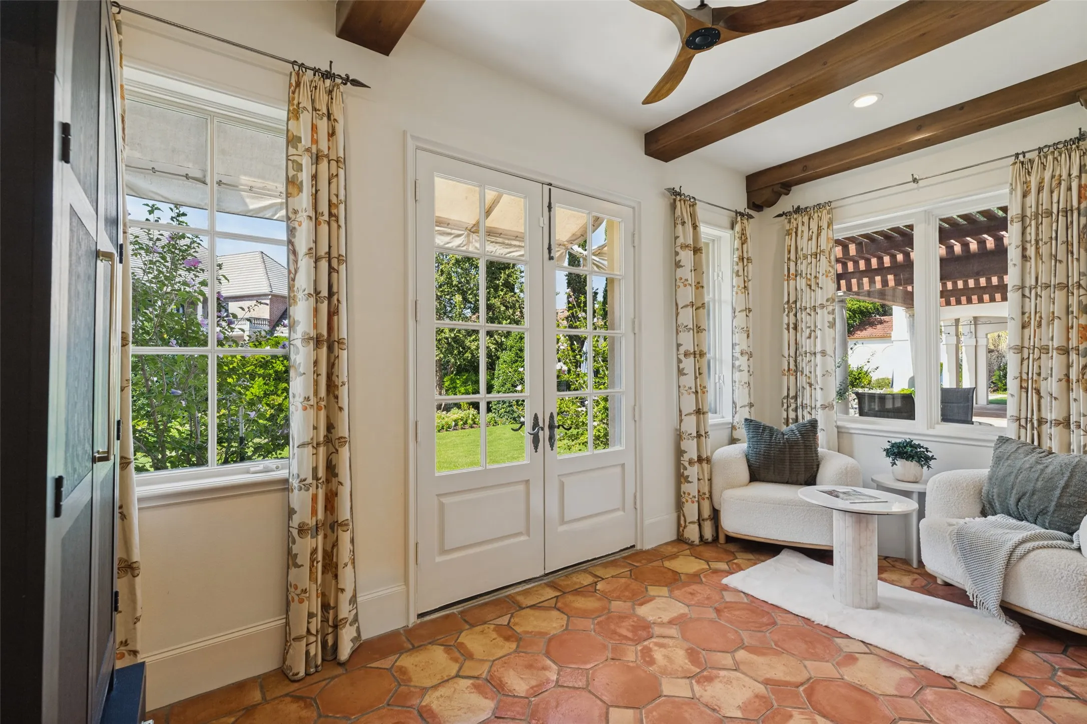 Exposed wood beam ceiling, wood-finish ceiling fan, terracotta tile flooring, white-framed French doors, and multiple white-framed windows
