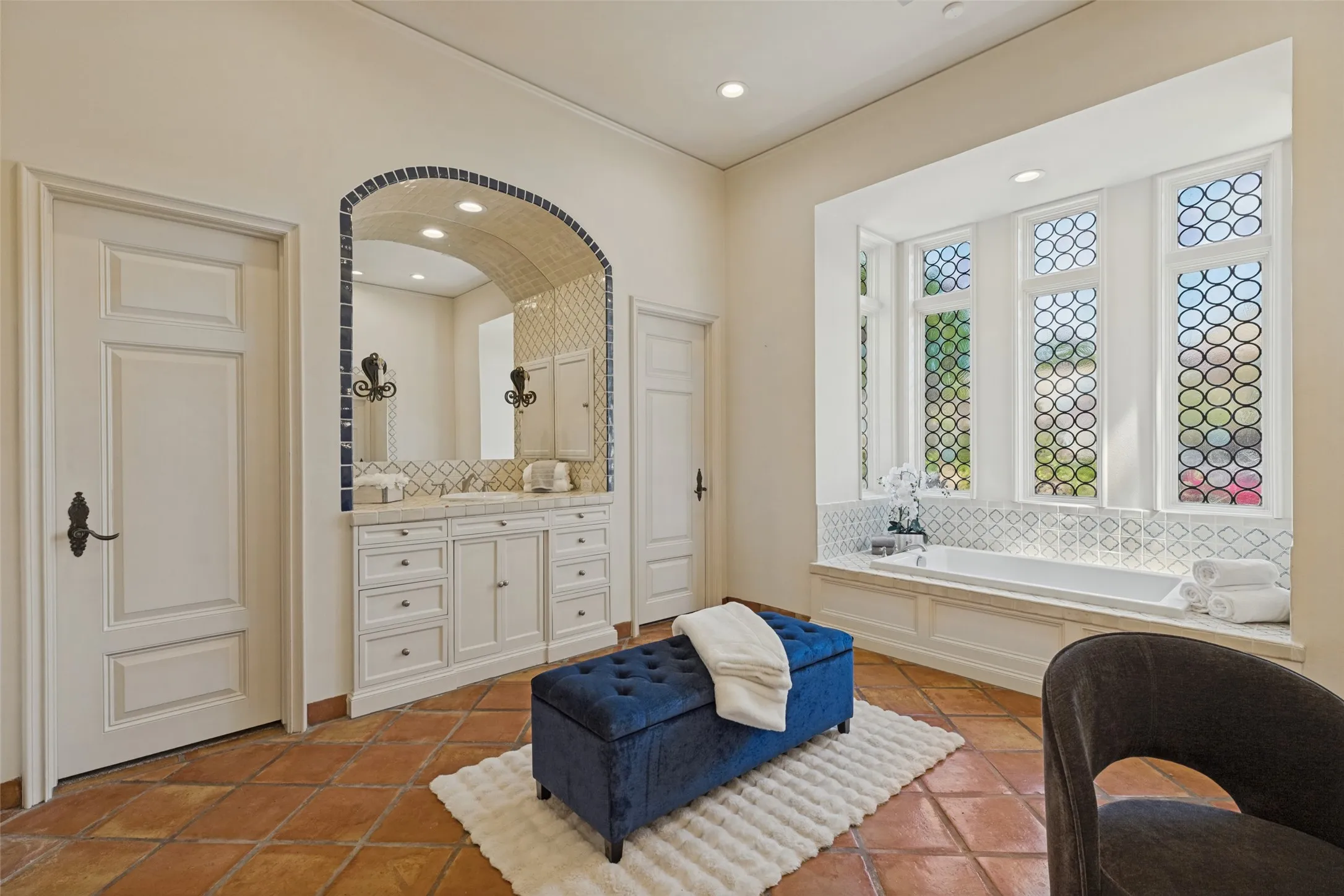 Built-in soaking tub with decorative tile surround, arched dual vanity with integrated mirror, and terracotta tile flooring