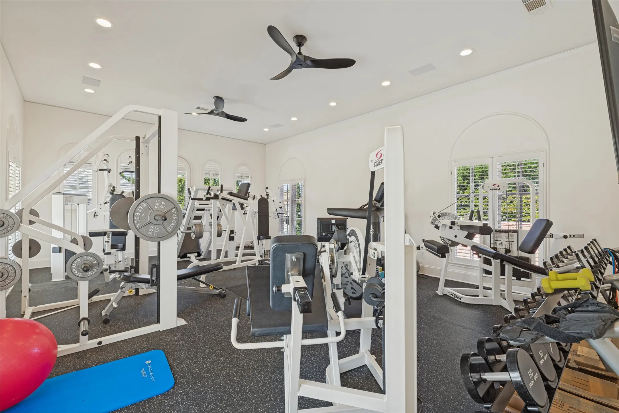Dedicated fitness room featuring recessed lighting, ceiling fans, arched windows with plantation shutters, and dark rubber flooring