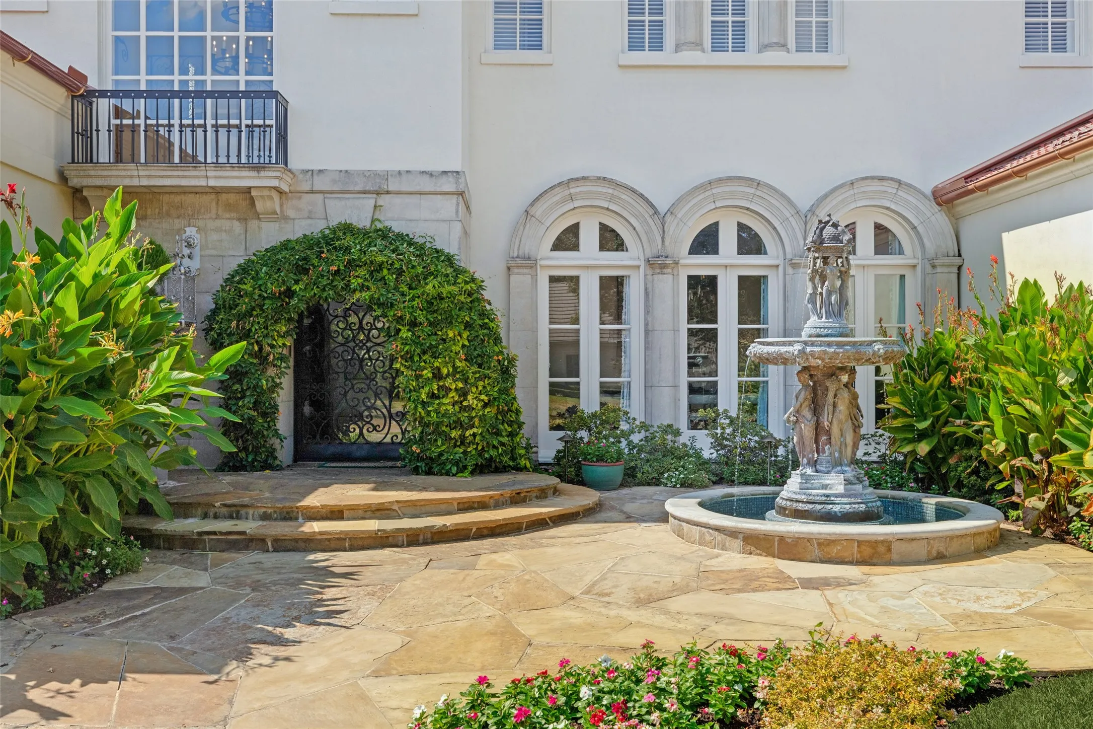 Stone patio with a tiered fountain, arched windows, and an ornamental iron gate covered in greenery
