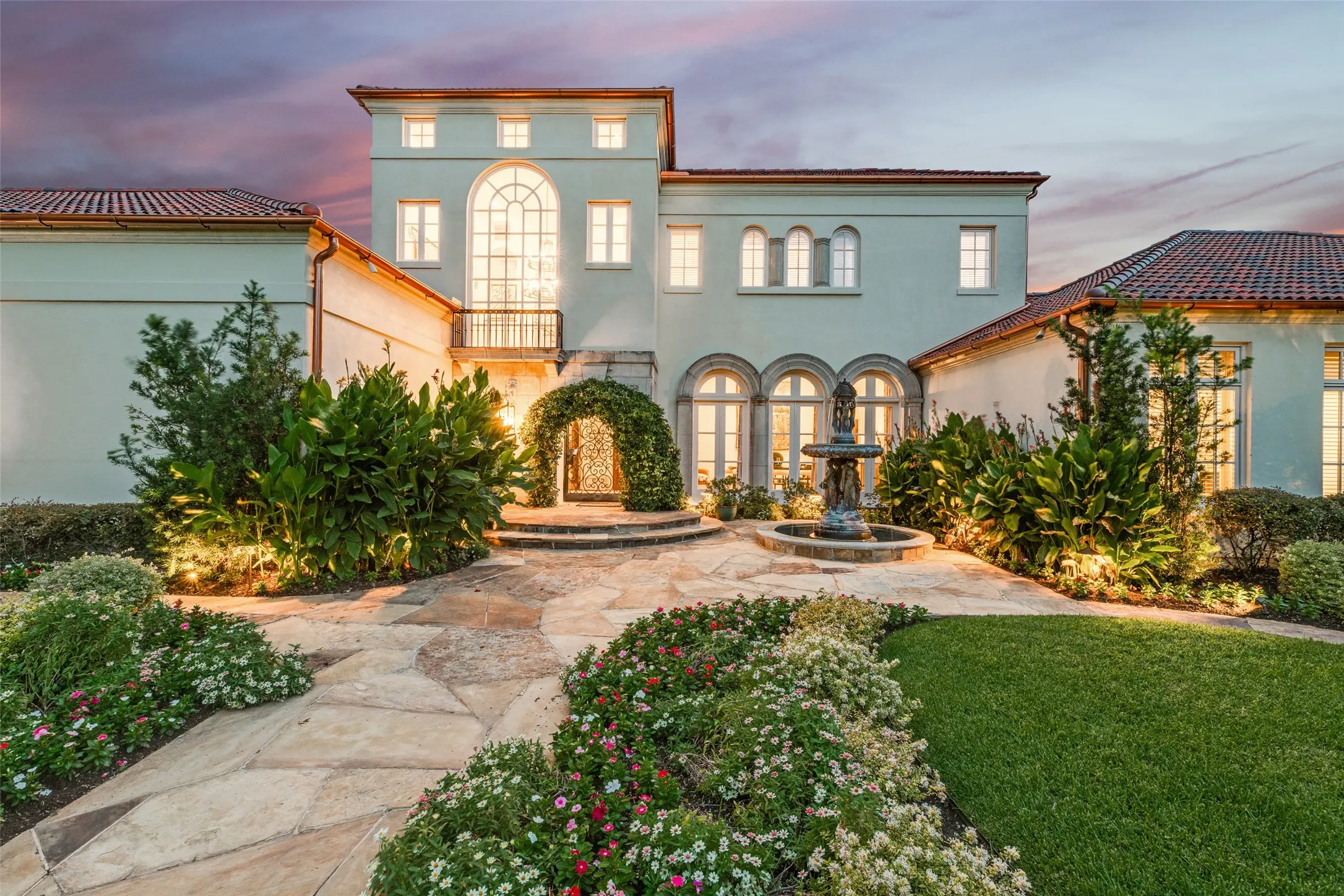 Stucco exterior with terracotta tile roofing, stone paver walkway, lush landscaping, and a tiered water fountain