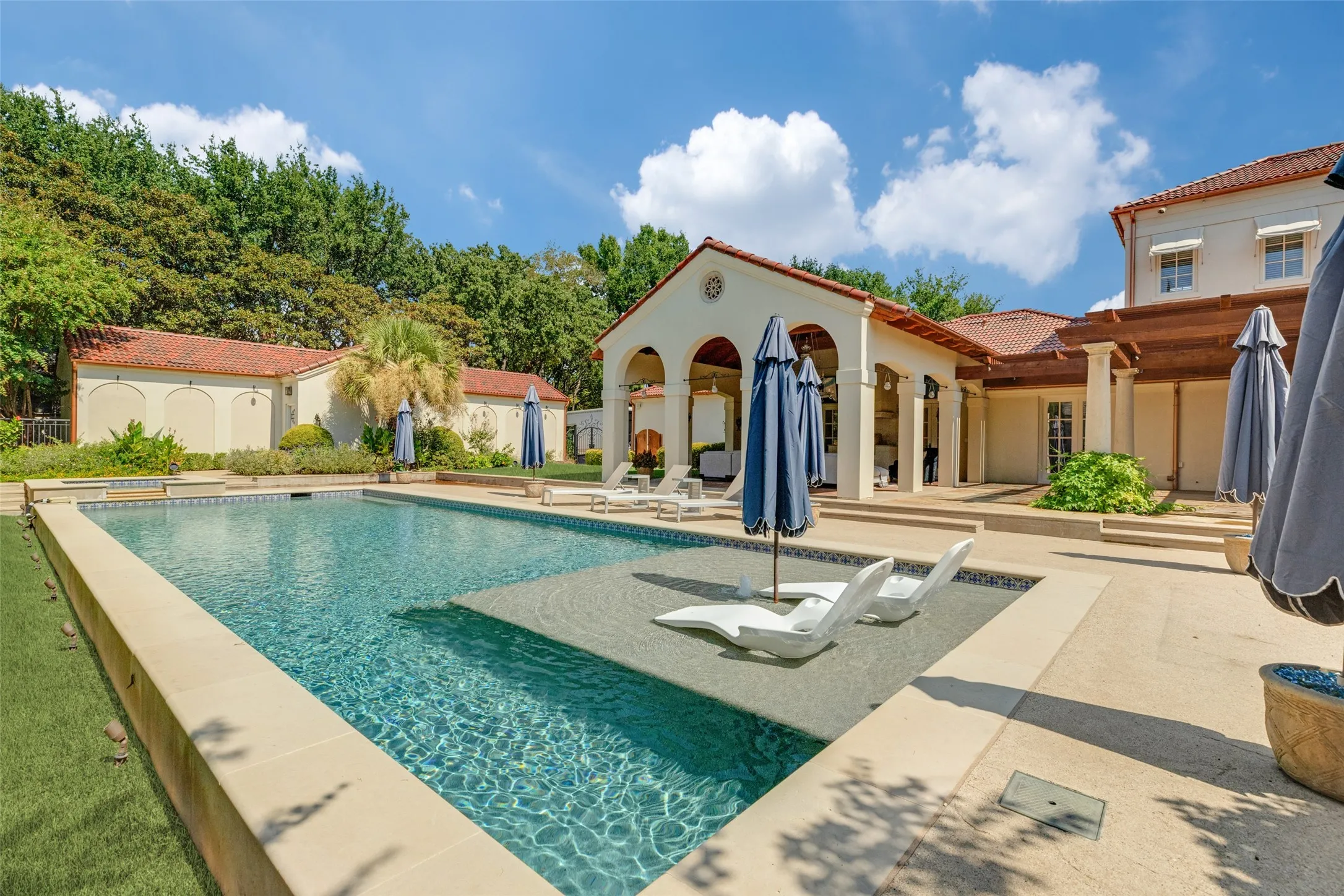 Expansive outdoor living area featuring a rectangular pool with an integrated sun shelf, surrounding stone coping, a covered patio with arched openings, and a red tile roof
