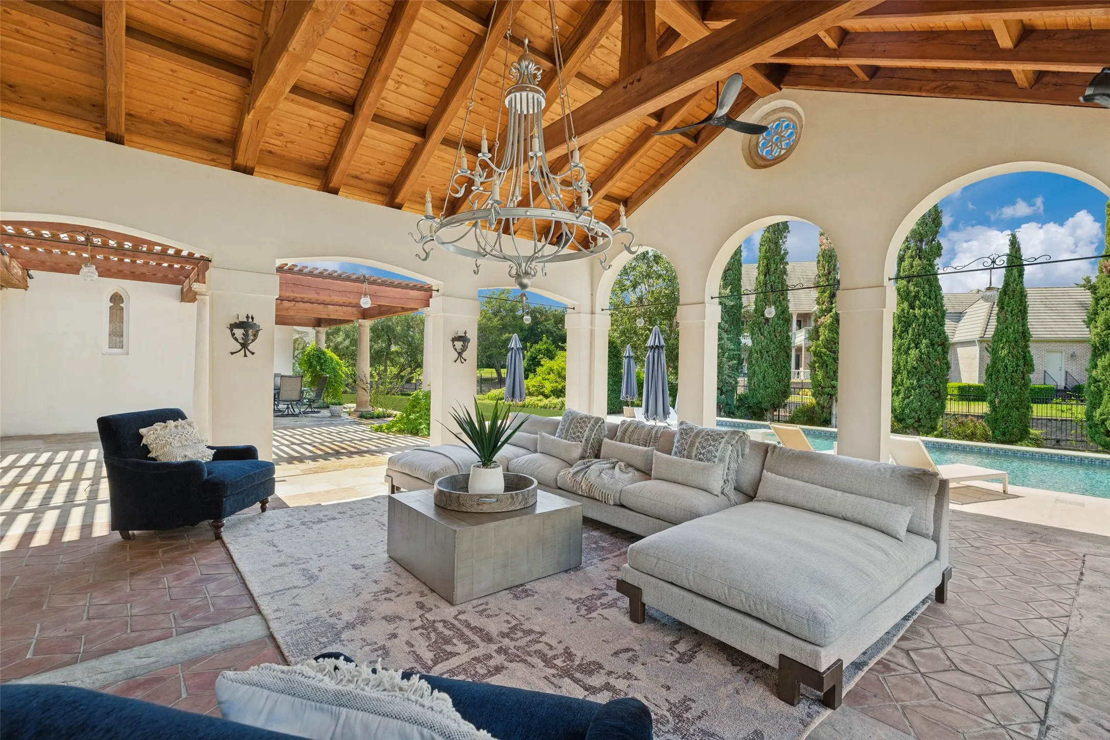 Covered outdoor living area with a wood-beamed ceiling, decorative chandelier, arched openings, and a paver stone patio