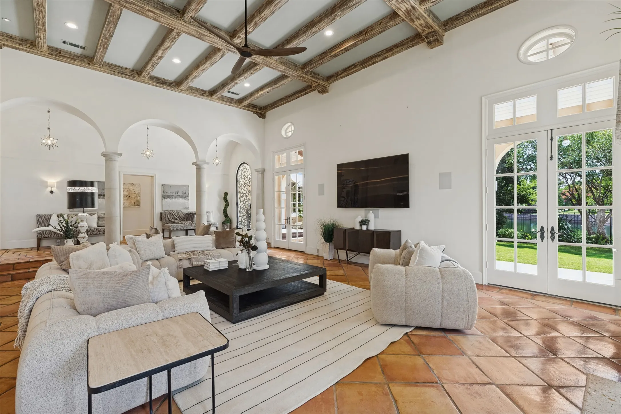 High ceilings with exposed wood beams, terracotta tile flooring, arched doorways, and white French doors with transom windows