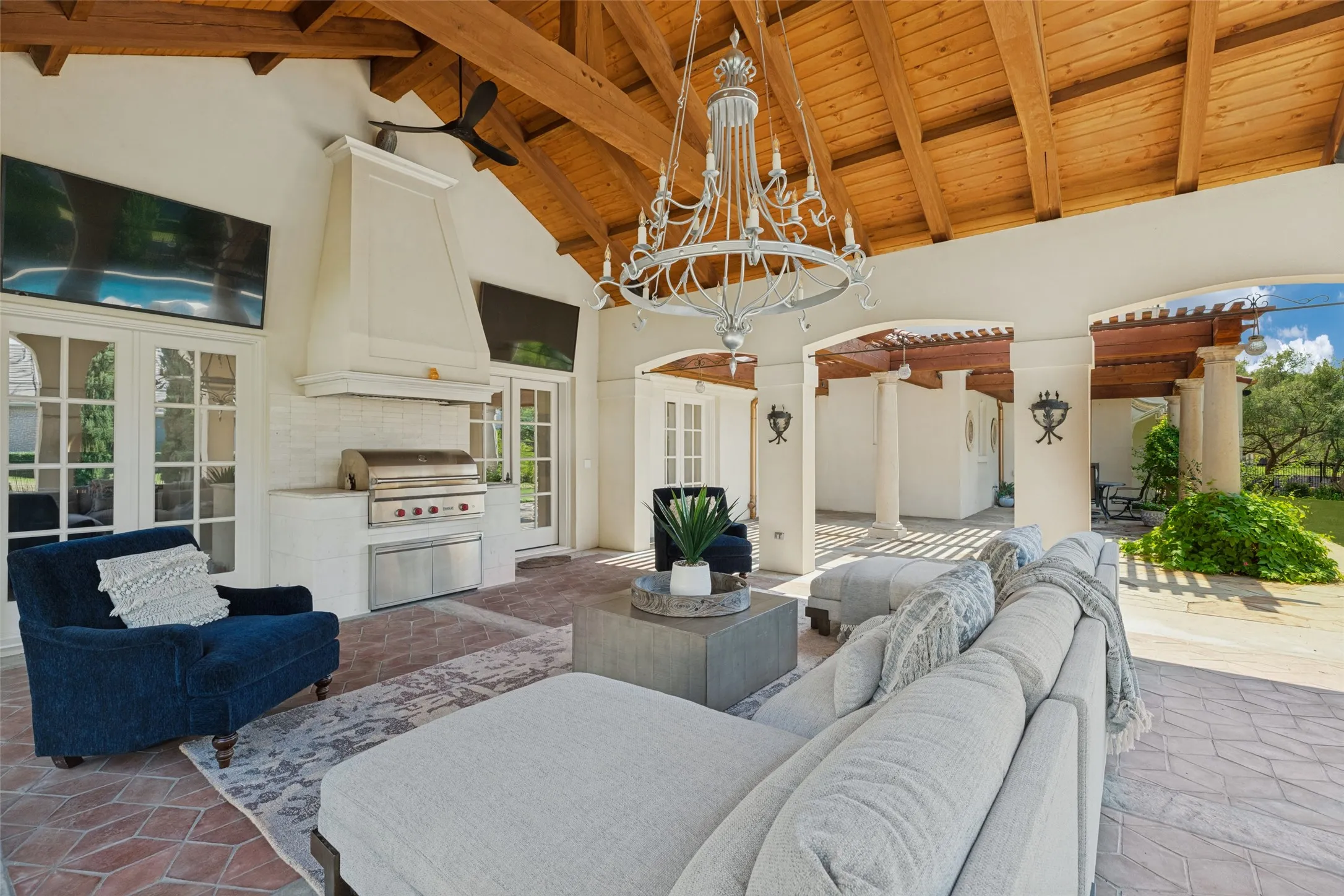 Vaulted wood-finish ceiling with exposed beams, integrated outdoor kitchen with built-in grill, white brick backsplash, French doors, and a decorative chandelier