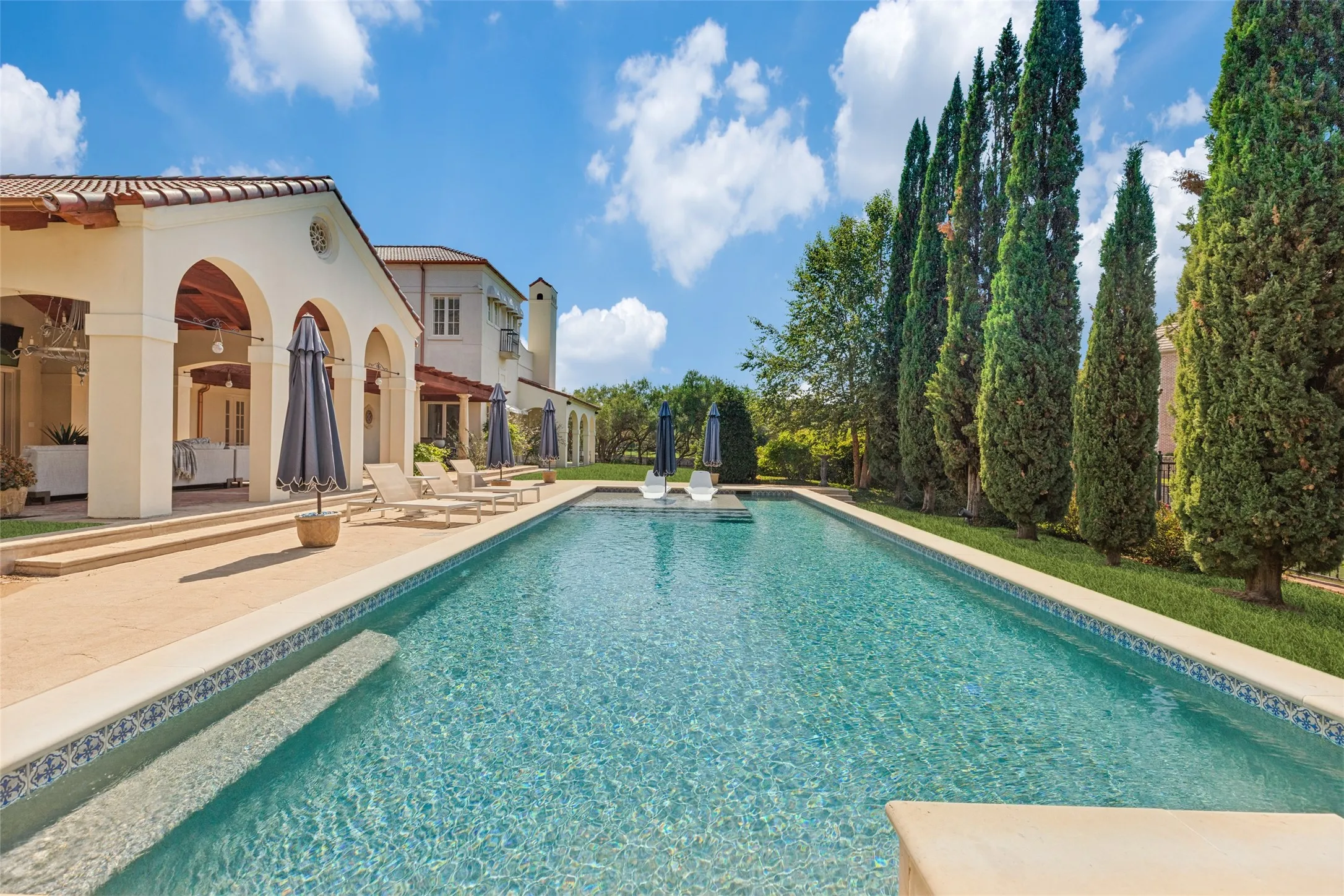 Elongated swimming pool with patterned tile border, expansive paver patio, arched exterior structure, and a row of tall, slender evergreen trees