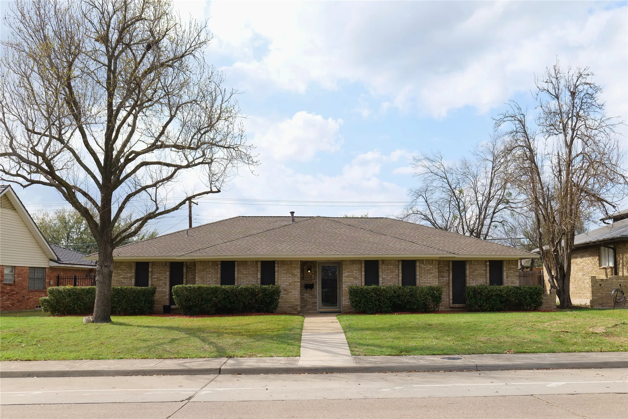 Brick exterior with a brown shingle roof, well-maintained lawn, mature trees, and a concrete walkway