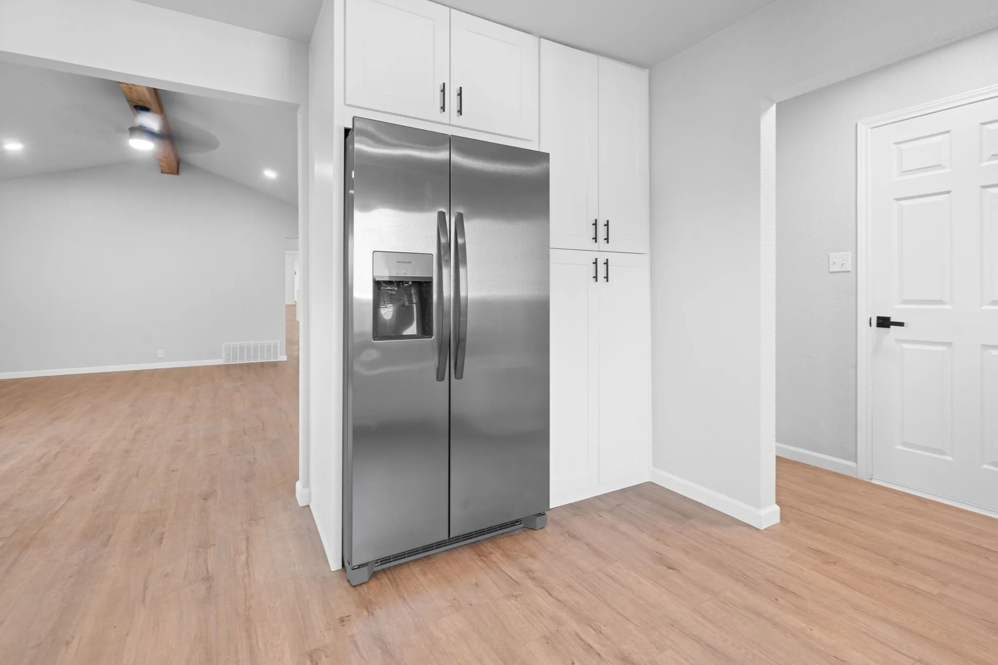 Stainless steel French door refrigerator, white shaker-style cabinetry with matte black hardware, wood-finish flooring, vaulted ceiling with exposed wood beam, recessed lighting