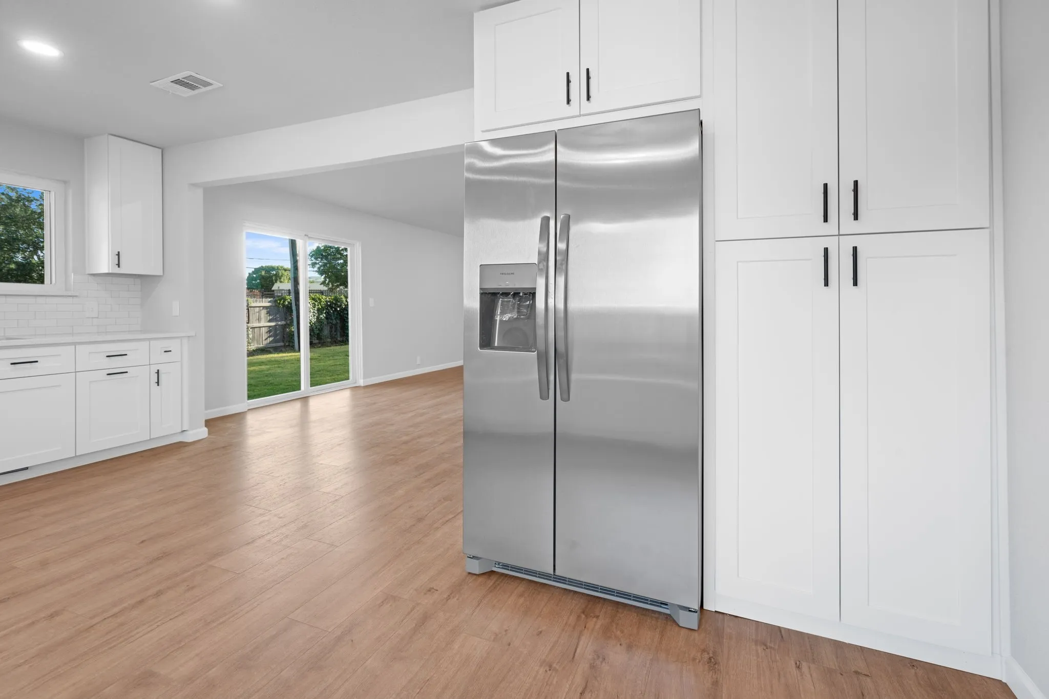 Stainless steel side-by-side refrigerator, white shaker-style cabinetry with matte black hardware, white subway tile backsplash, wood-finish flooring, and sliding glass patio doors