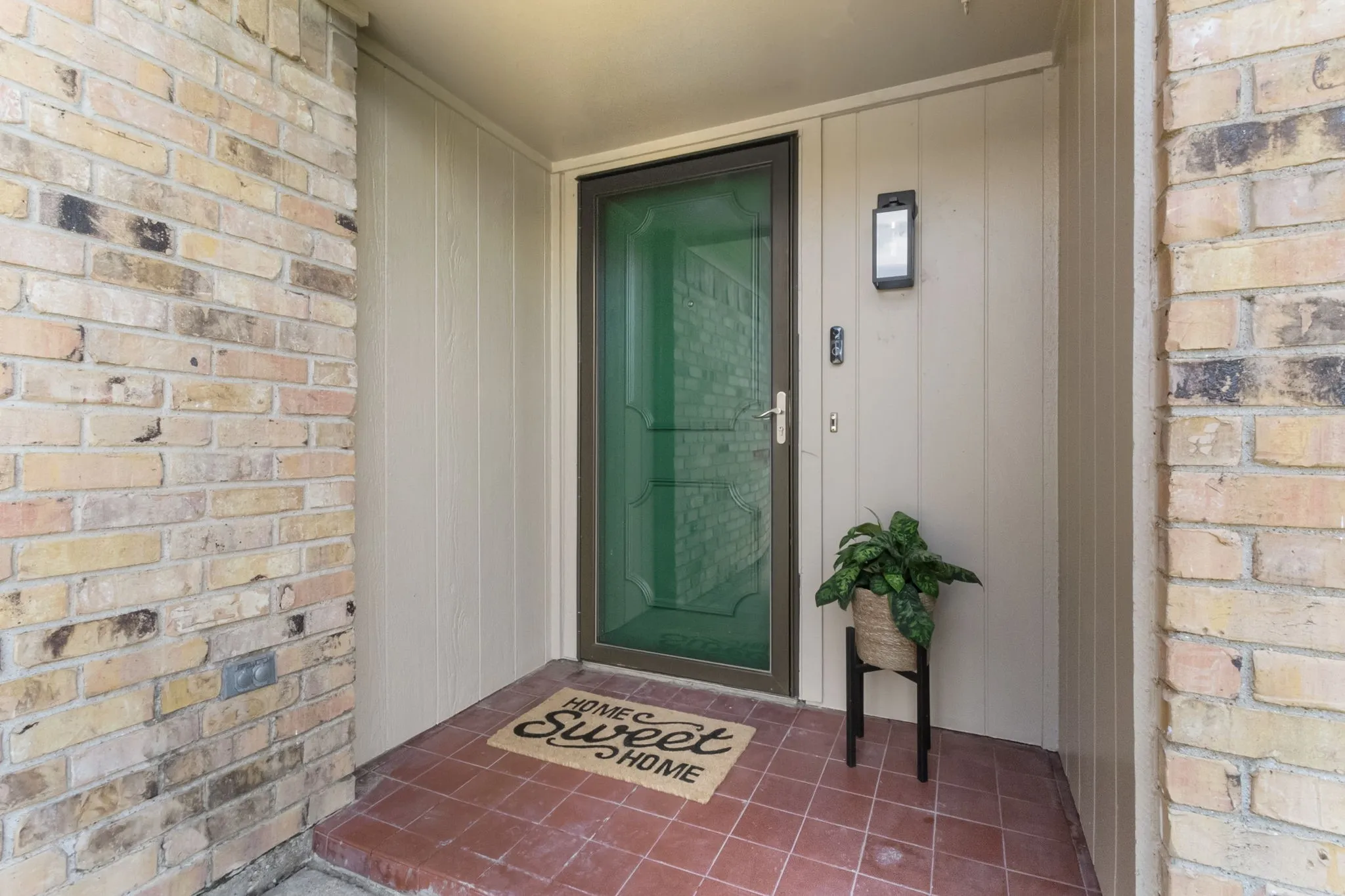 Brick and vertical panel siding exterior with a security door, terracotta tile flooring, and an outdoor wall sconce