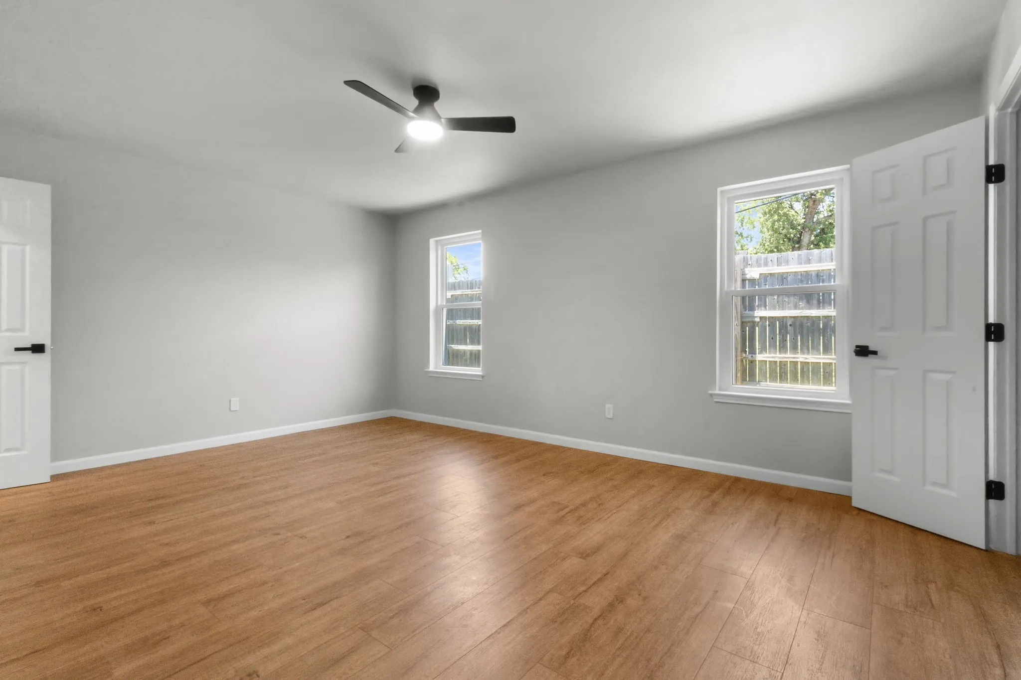 Spacious room featuring wood-finish flooring, light grey painted walls, ceiling fan, and two panel doors with matte black hardware