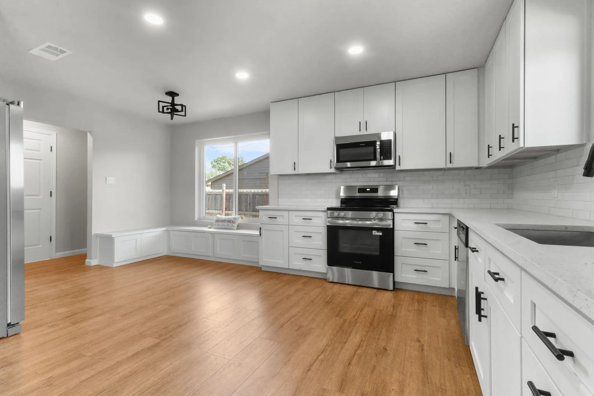 Wood-finish flooring, white cabinetry with black hardware, stainless steel appliances, white subway tile backsplash, and a built-in window bench