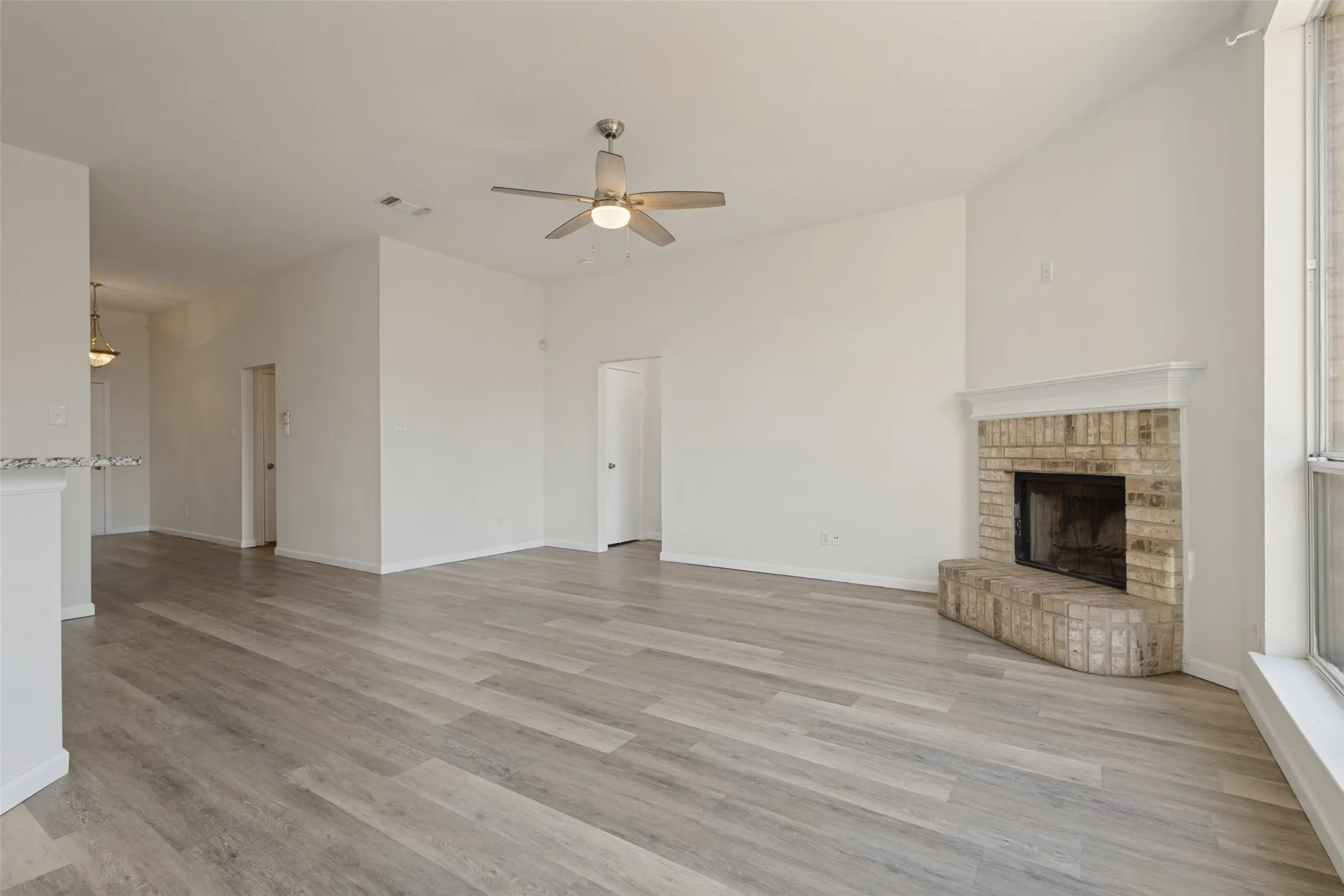 Open-concept living area featuring wood-finish flooring throughout, a ceiling fan with integrated lighting, a brick fireplace with stone surround, and abundant natural light from large windows