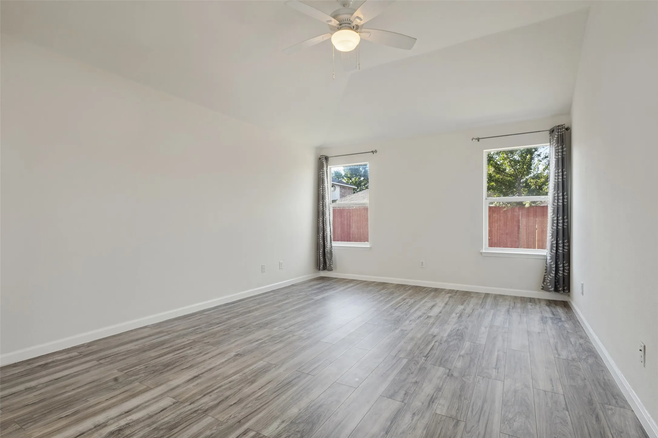 Vaulted ceiling with a ceiling fan, two windows, wood-finish flooring, and light-toned walls