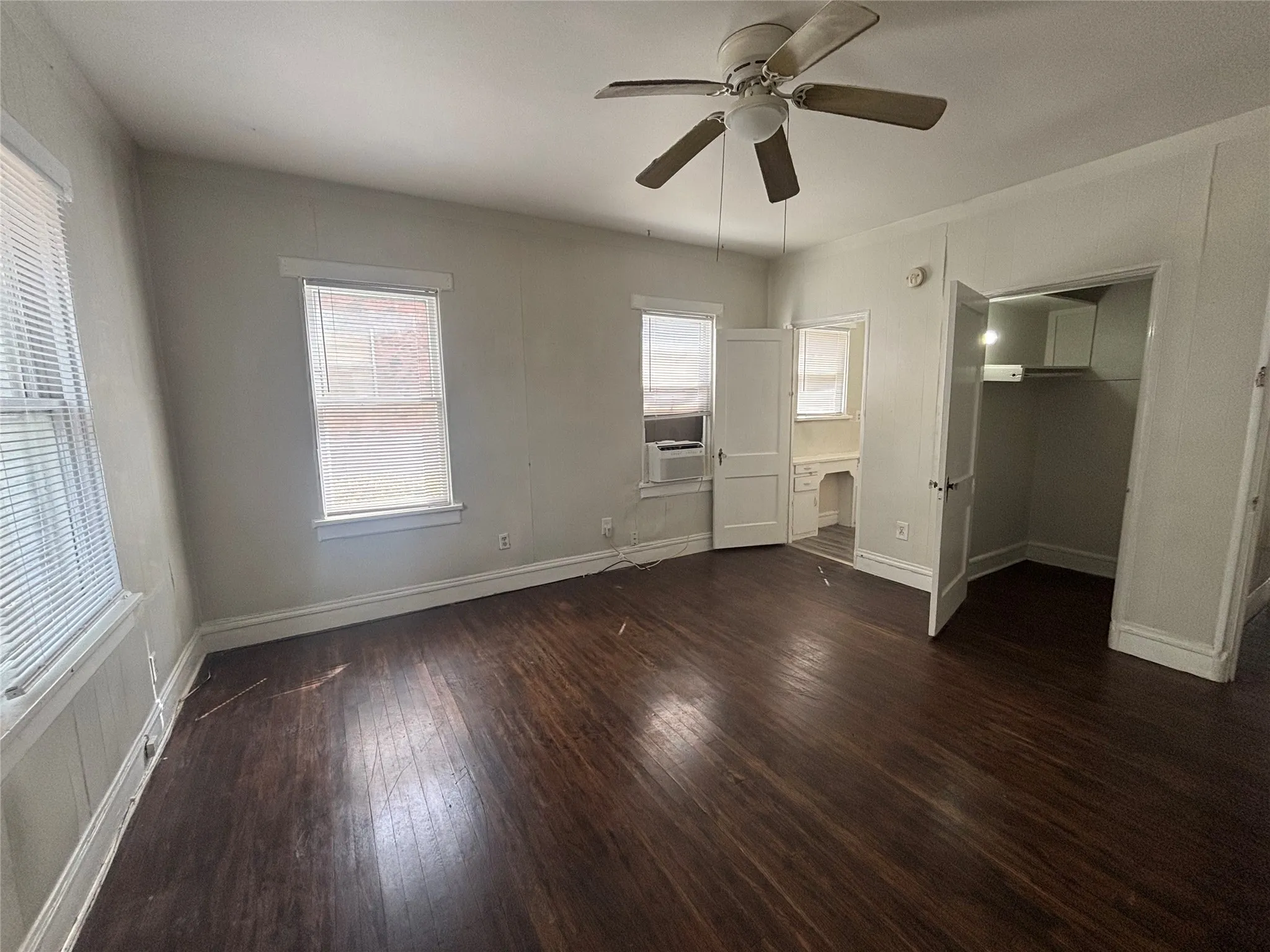 Dark wood-finish flooring, ceiling fan with integrated lighting, multiple windows, built-in closet, and an in-wall air conditioning unit