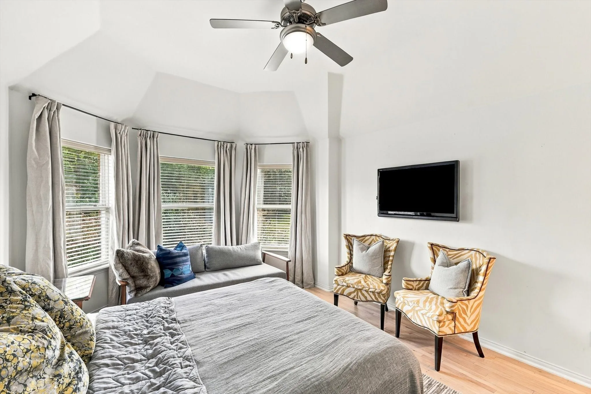 Light wood-finish flooring, vaulted ceiling, ceiling fan with integrated lighting, and multiple windows with blinds
