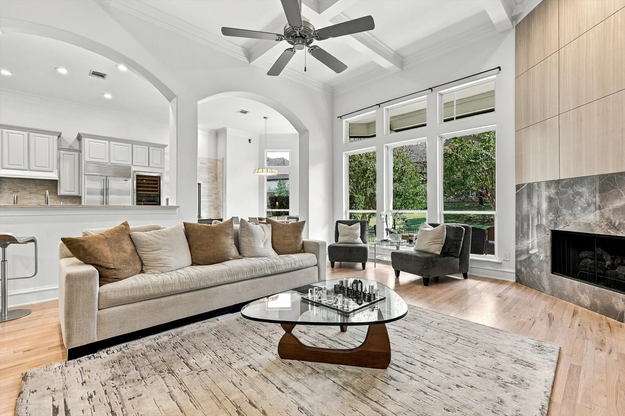 Hardwood flooring, floor-to-ceiling windows, stone fireplace surround, coffered ceiling with a ceiling fan, and arched room openings