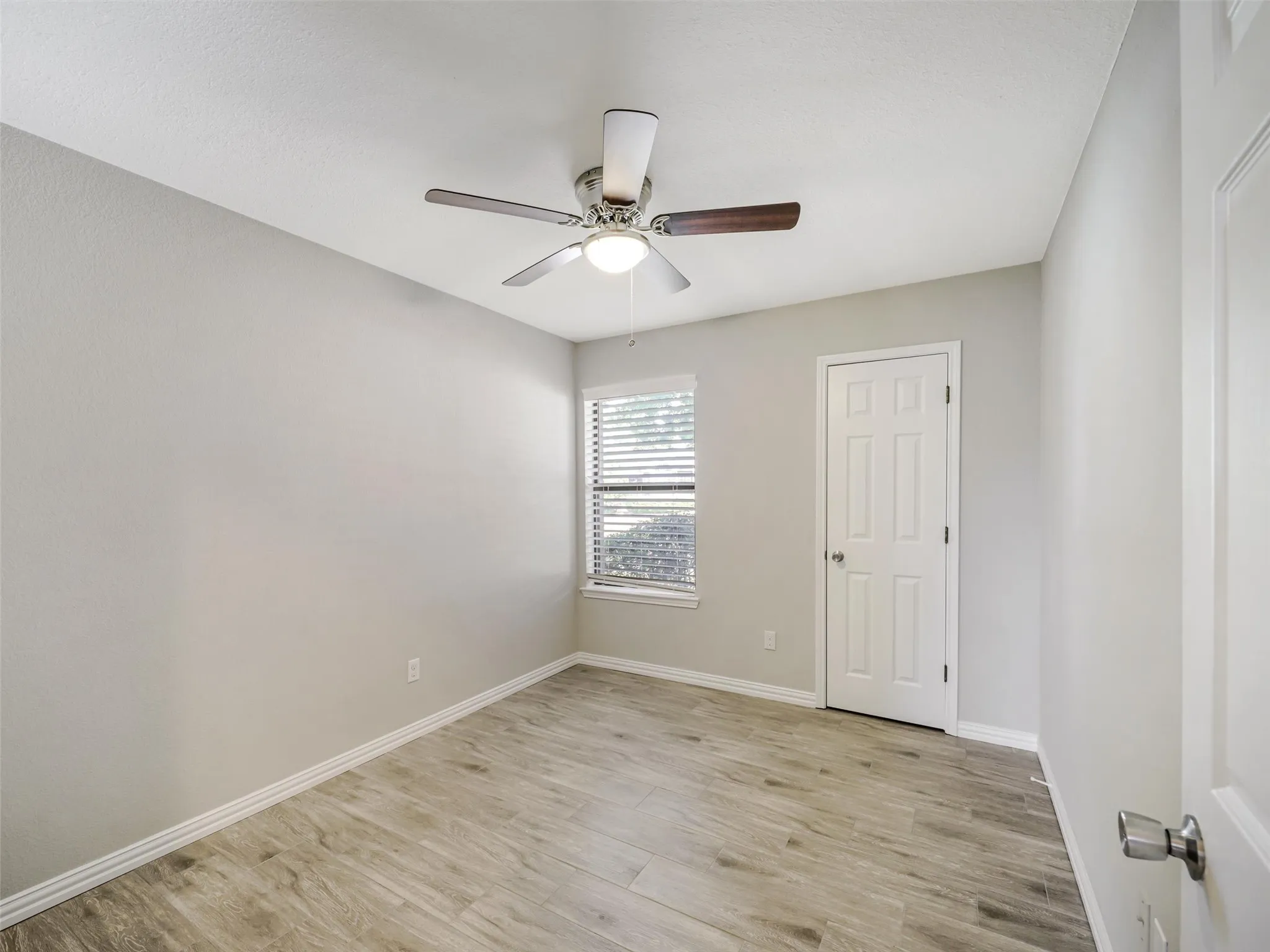 Neutral wall color, wood-finish flooring, ceiling fan with integrated lighting, window with horizontal blinds, and a paneled door