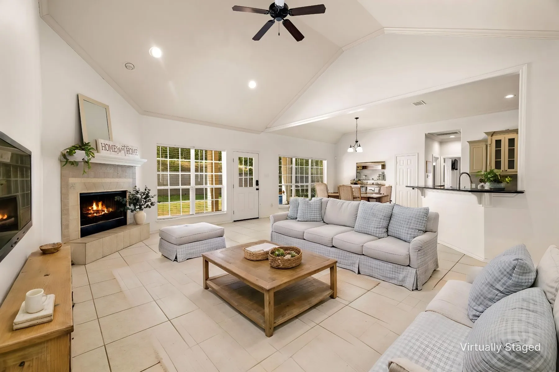 Vaulted ceilings, a stone-clad fireplace, large grid-pane windows, light tile flooring, and an open-concept pass-through kitchen