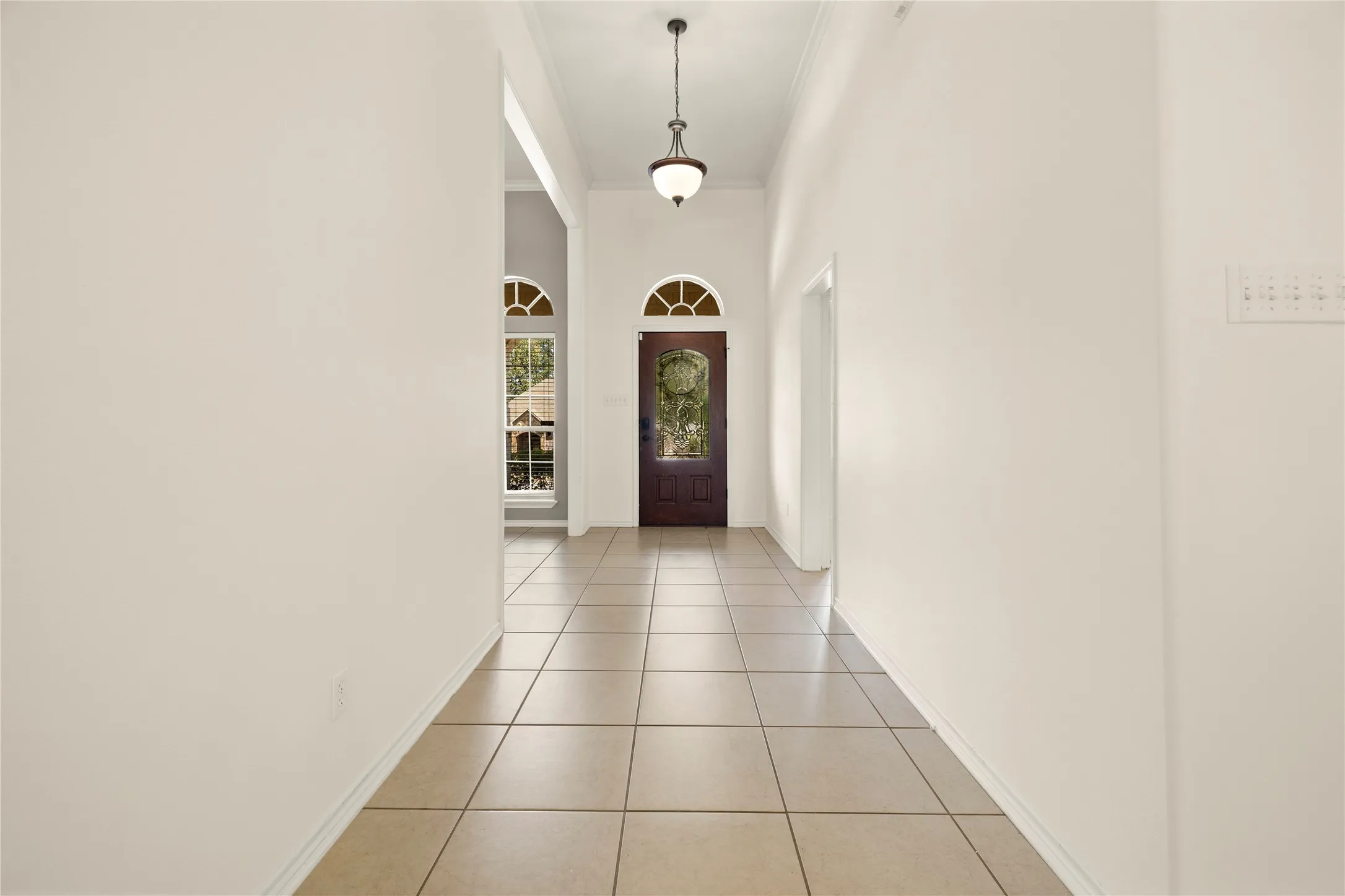 Tiled flooring extending through a hallway with arched doorways, a dark wood-finish door with an arched transom window, and a decorative pendant light fixture