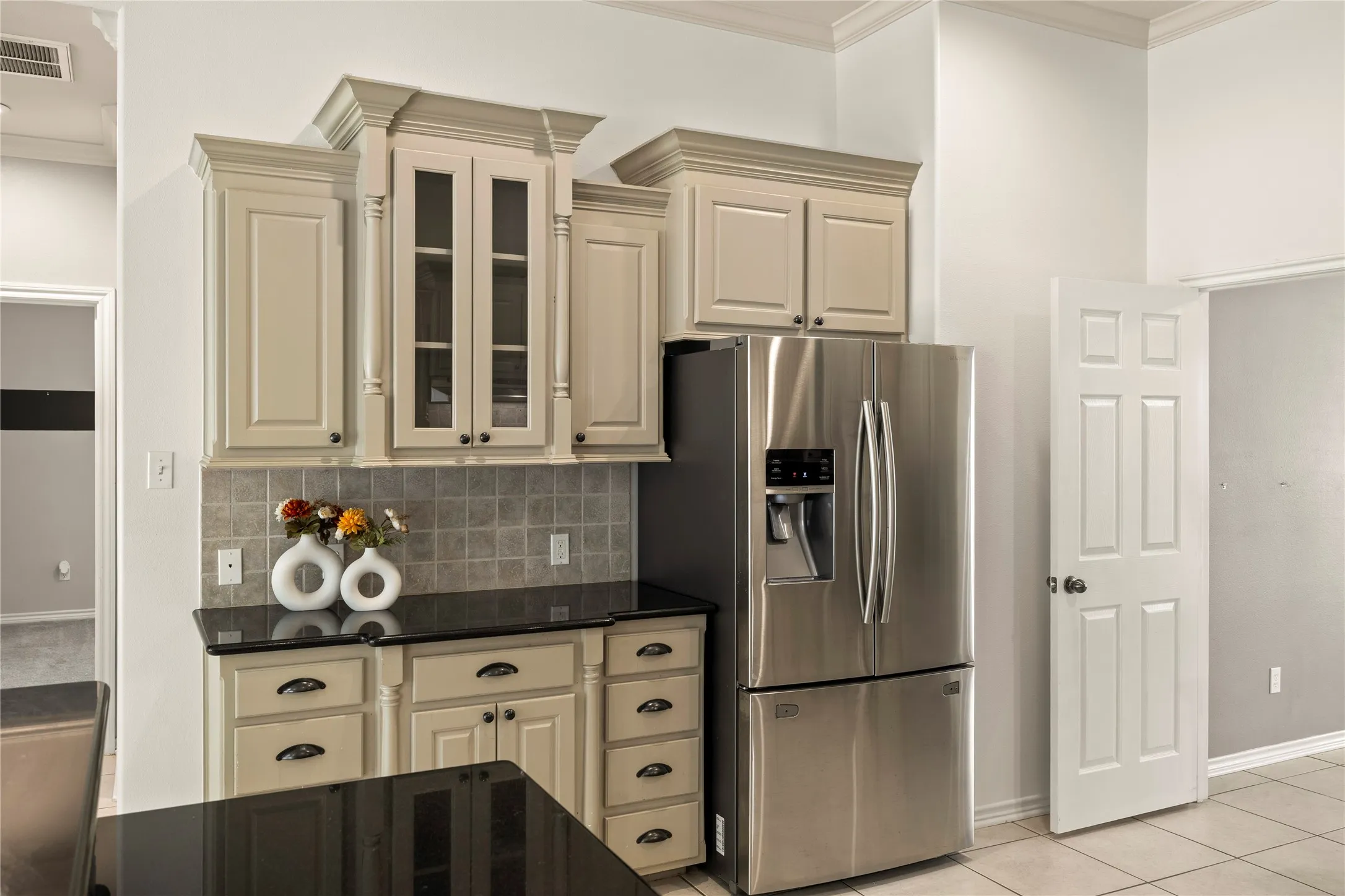 Built-in stainless steel refrigerator, light-toned cabinetry with crown molding, glass-front upper cabinets, dark countertops, and tiled backsplash