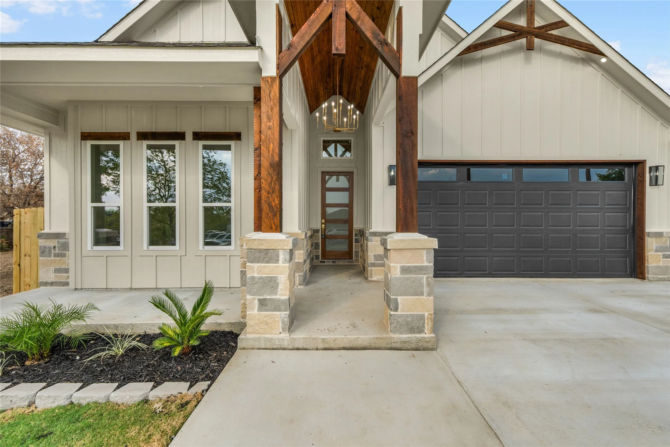 Board and batten siding with stone accents, exposed wood beams, a dark wood entry door with frosted glass, a decorative chandelier, and an attached two-car garage