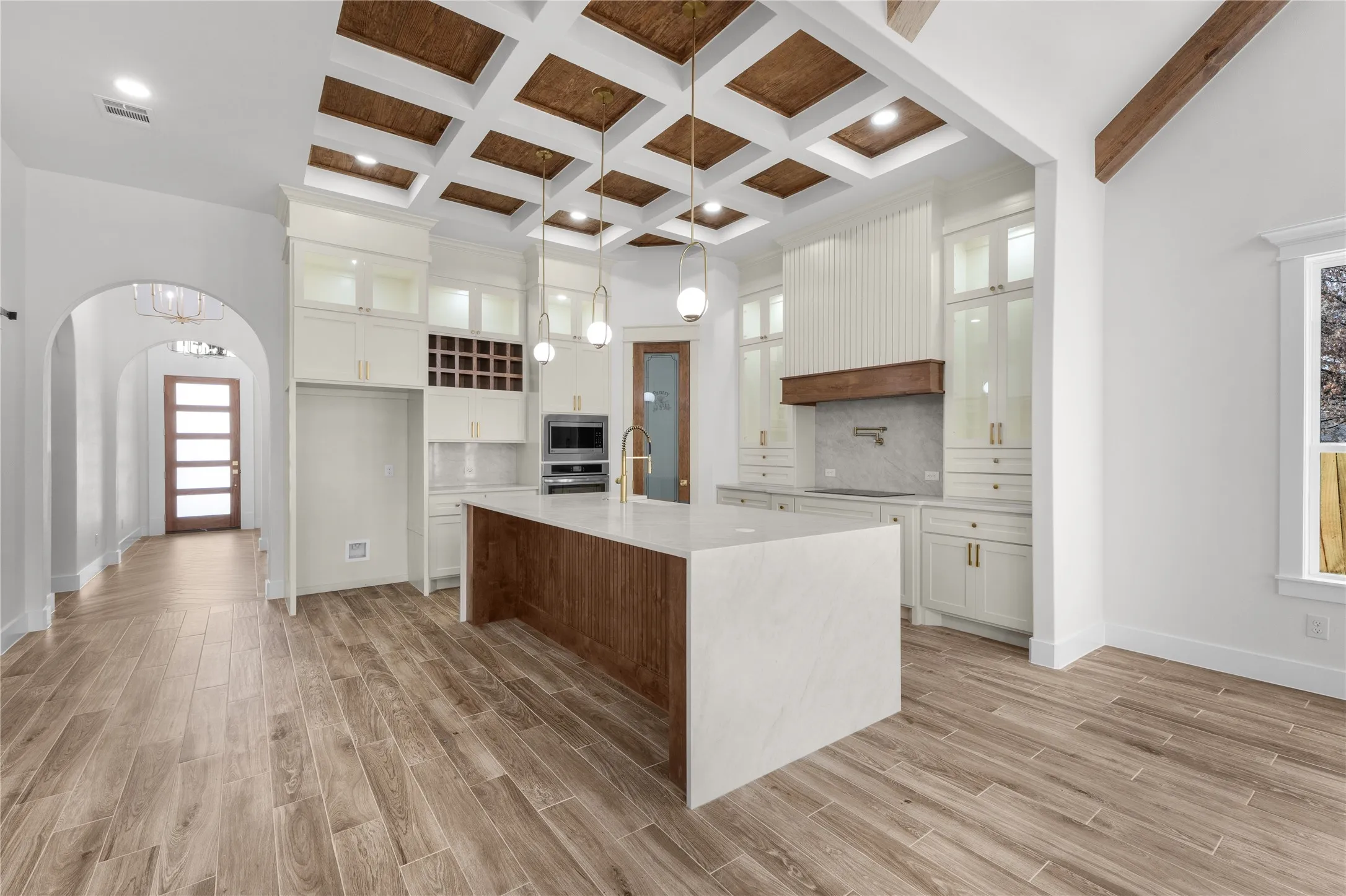 Coffered ceiling with wood-finish inserts, wood-finish flooring, kitchen island with waterfall countertop, built-in stainless steel appliances, and white cabinetry