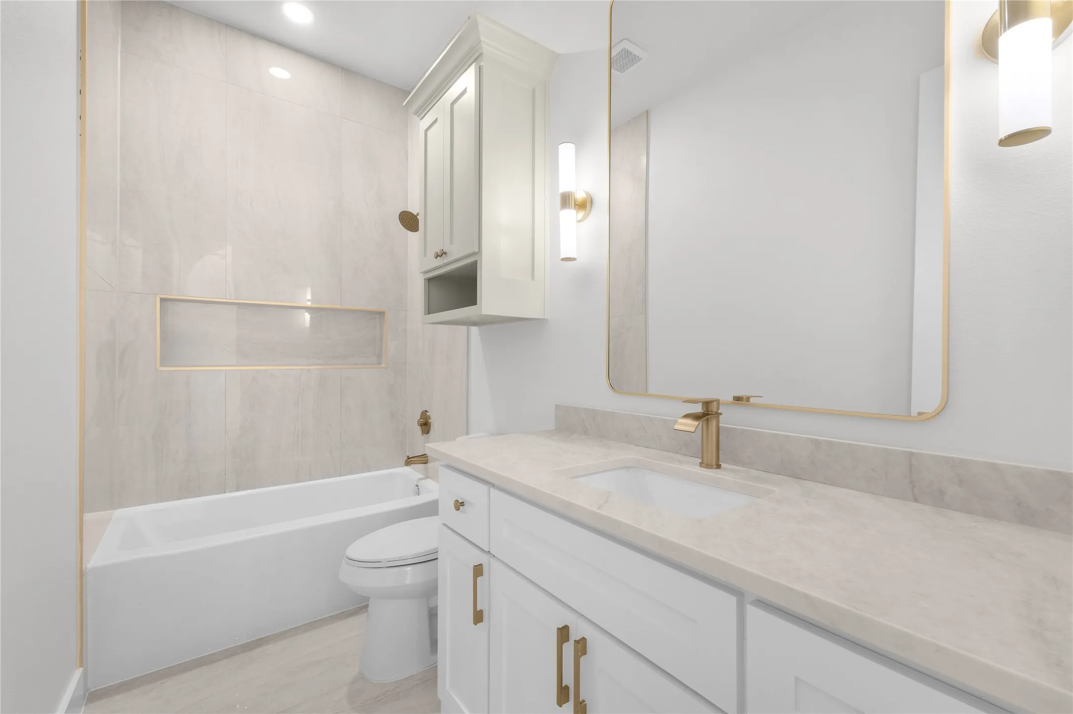 Bathtub with tiled surround and integrated niche, white vanity with gold-tone hardware, undermount sink, and stone-finish countertop, framed mirror with integrated lighting
