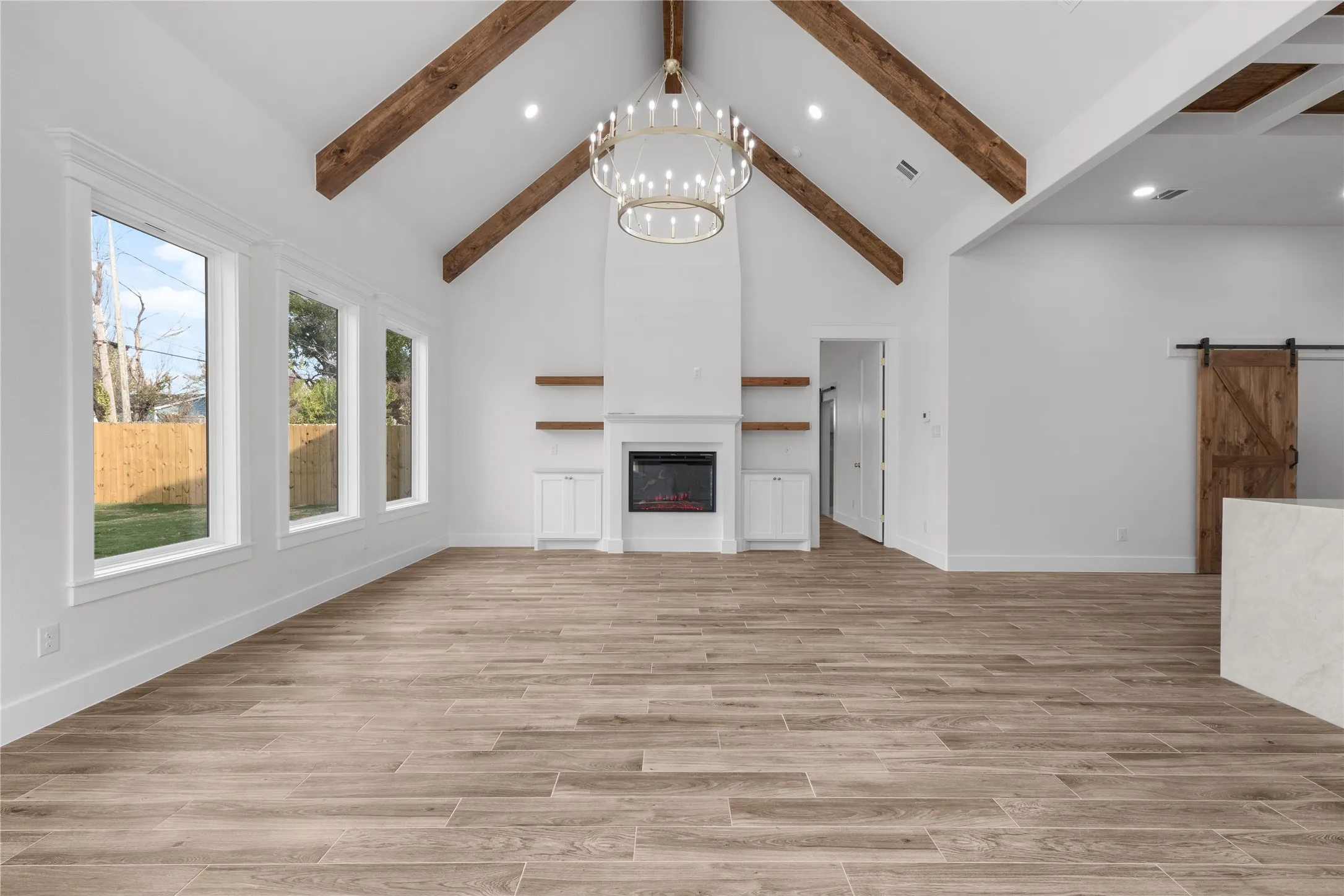 Vaulted ceiling with exposed wood beams, wood-finish flooring, built-in electric fireplace, multiple windows with white trim, and a decorative chandelier