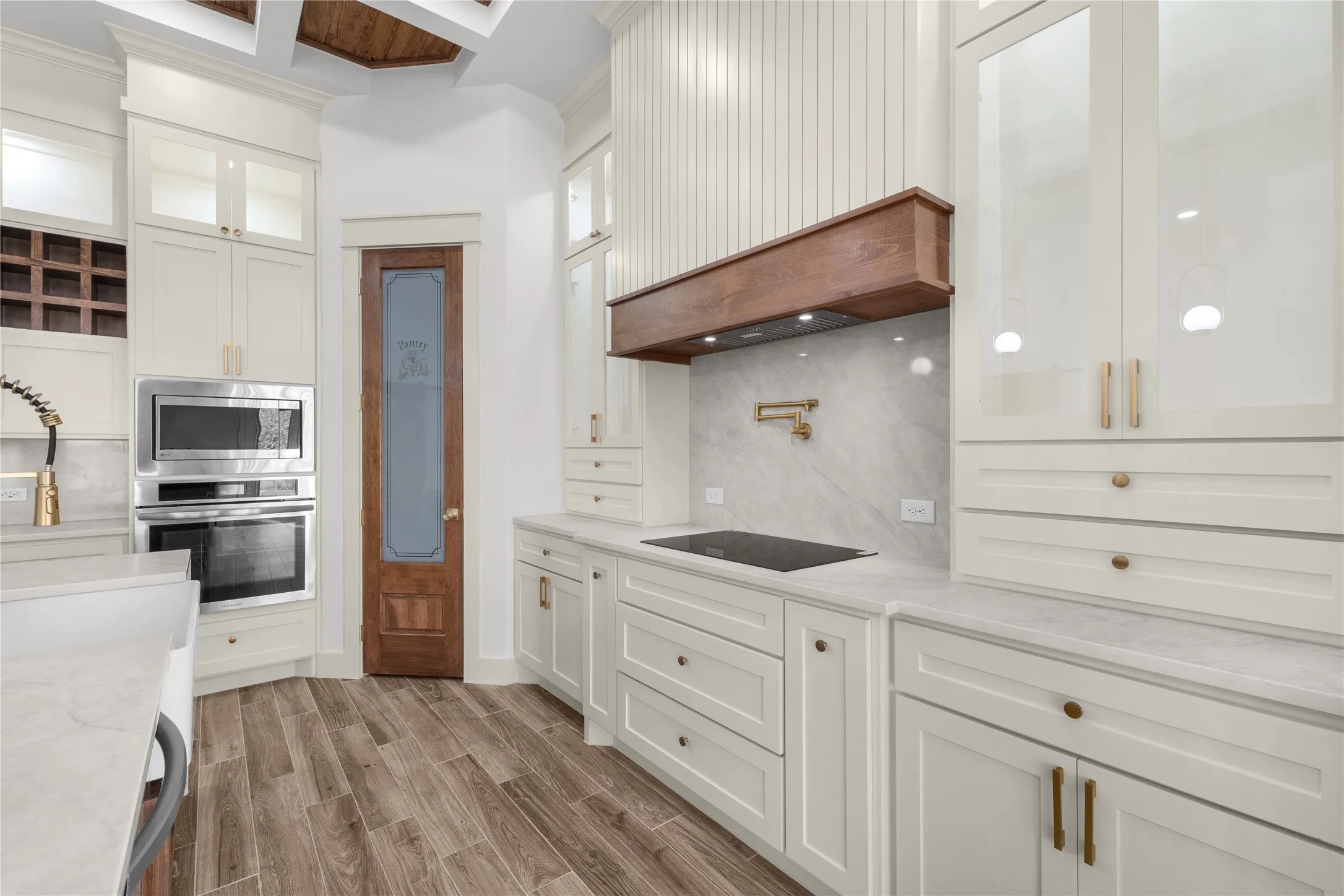 White cabinetry with gold-toned hardware, stainless steel double ovens, wood-finish flooring, a pot filler, and a visible range