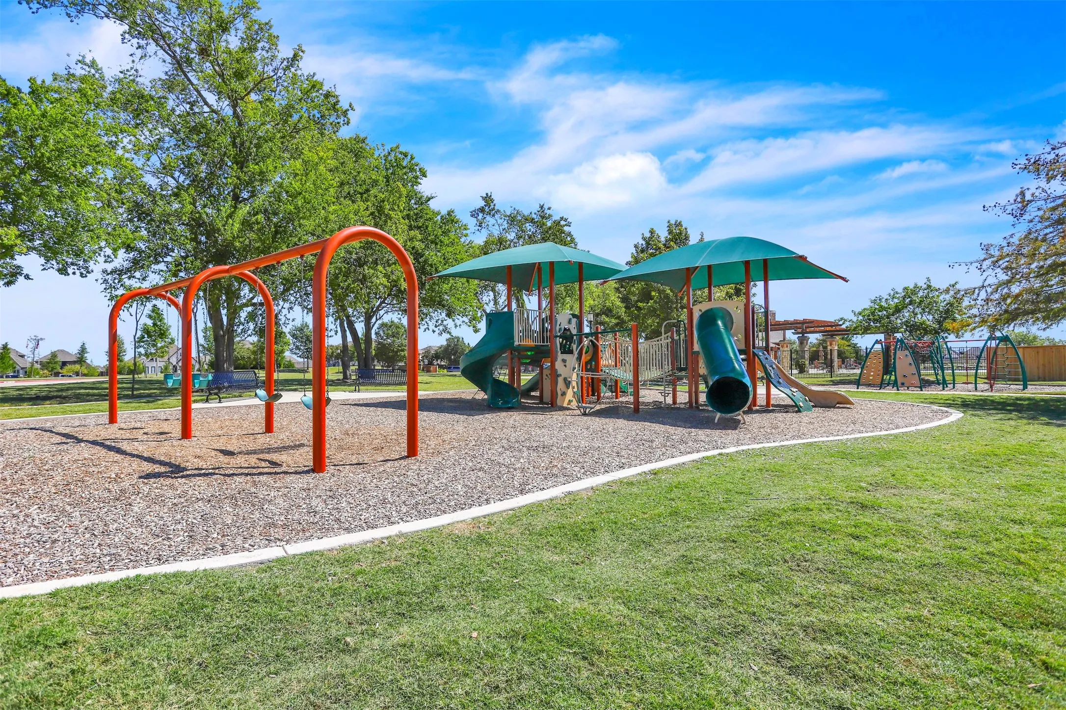 Expansive park area featuring a swing set, multiple play structures with slides, shade canopies, and a wood chip surface