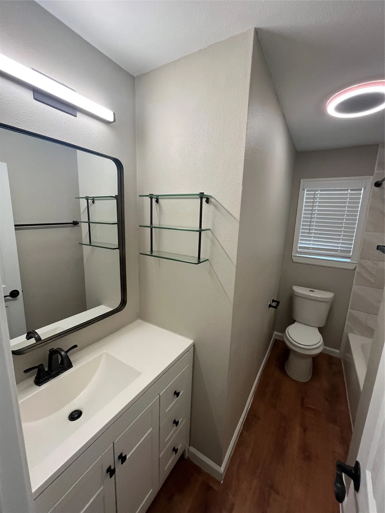 White vanity with integrated sink, black hardware, wood-finish flooring, and a contemporary wall-mounted light fixture