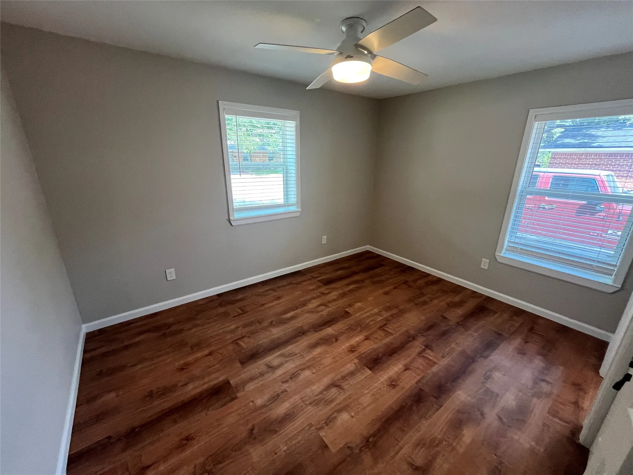 Wood-finish flooring, neutral wall color, two windows with blinds, and a ceiling fan with integrated lighting
