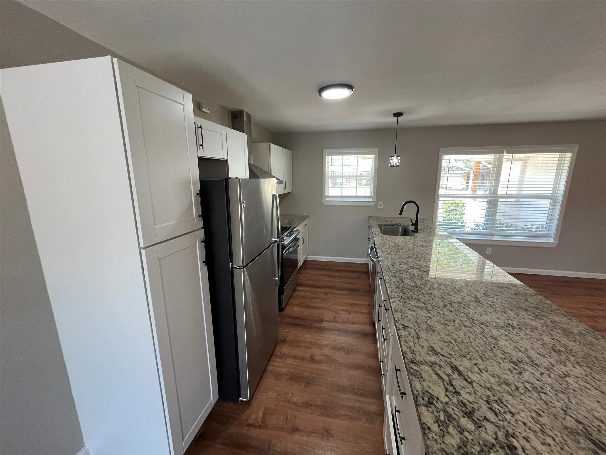 Expansive kitchen island with granite countertop, stainless steel appliances, white shaker-style cabinetry, wood-finish flooring, and recessed lighting