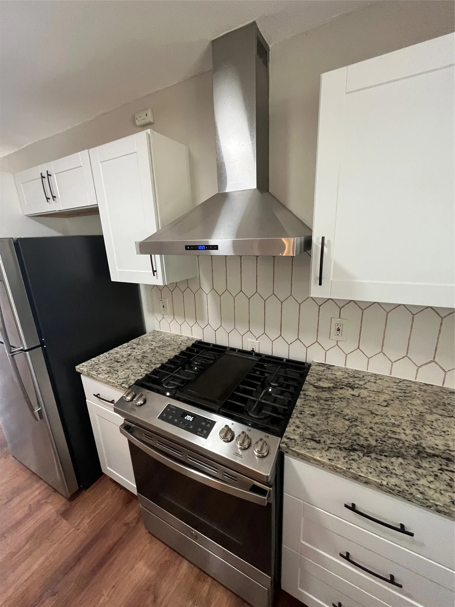 Stainless steel range hood and appliances, white shaker-style cabinetry, granite countertops, geometric tile backsplash, and wood-finish flooring