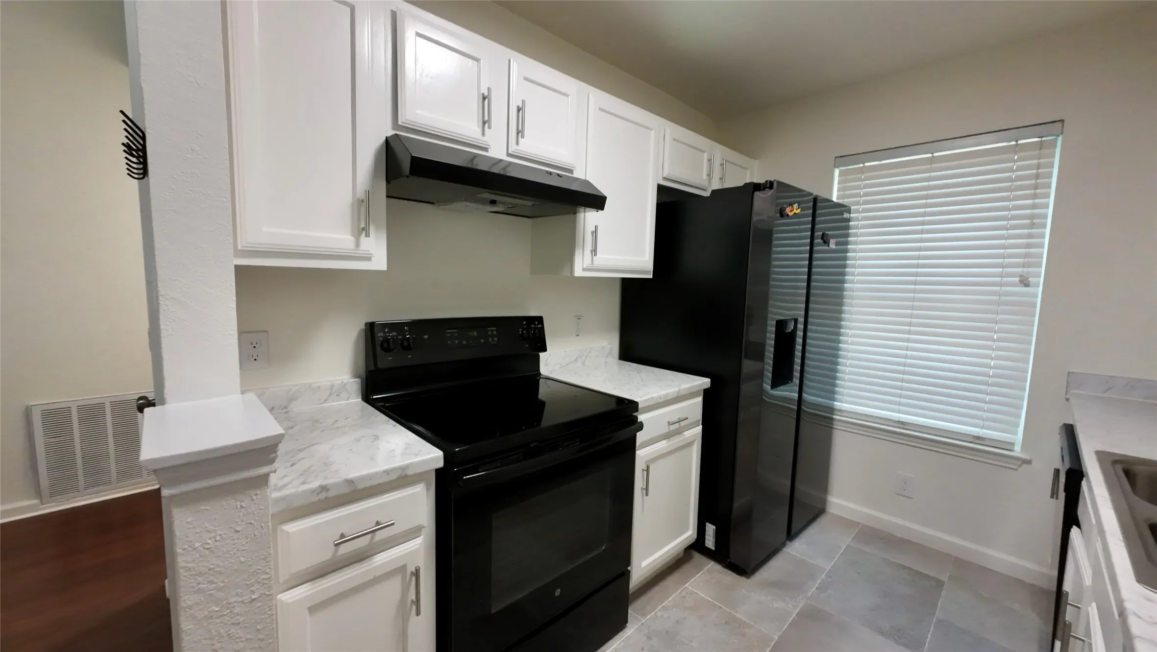 White cabinetry, light-toned countertops, black appliances, window with blinds, and tiled flooring