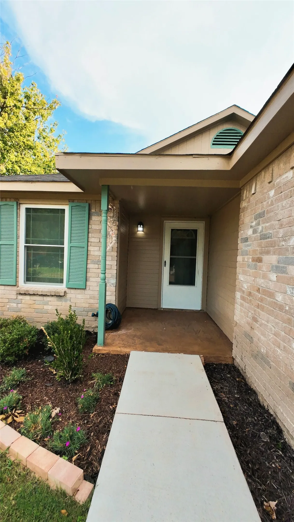 Brick exterior with mint green shutters and a covered entry, featuring a concrete walkway and landscaped planting beds