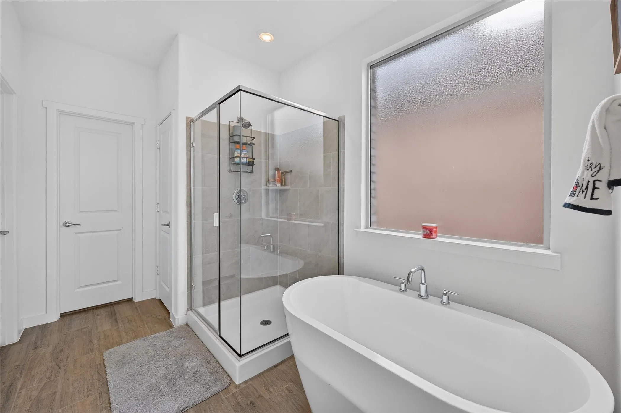 Freestanding soaking tub with chrome fixtures, glass-enclosed shower with tile surround, and wood-finish flooring