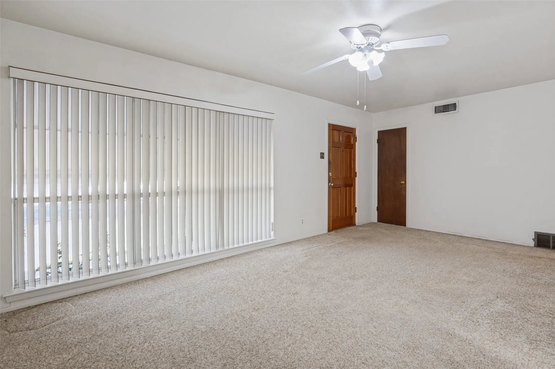 Spacious room with light-colored carpet, white walls, and a large window with vertical blinds