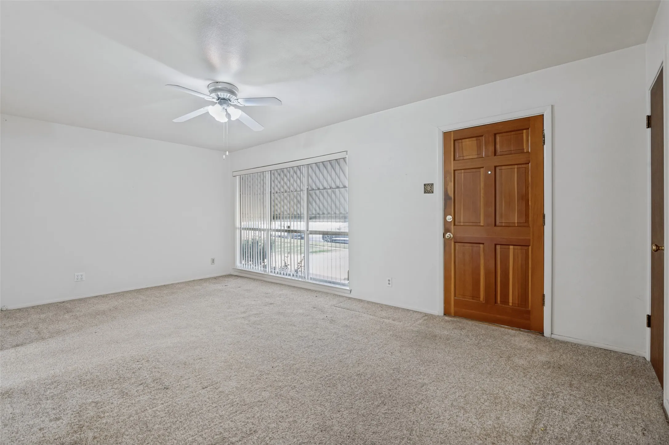 Carpeted interior with a paneled wood entry door, white ceiling fan, and large window with blinds