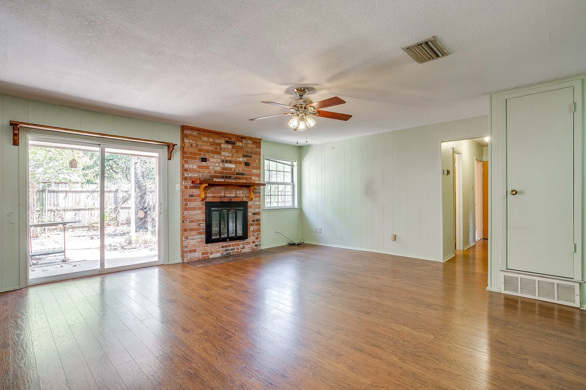 Wood-finish flooring, brick fireplace with mantel, sliding glass door to exterior, ceiling fan, and paneled walls
