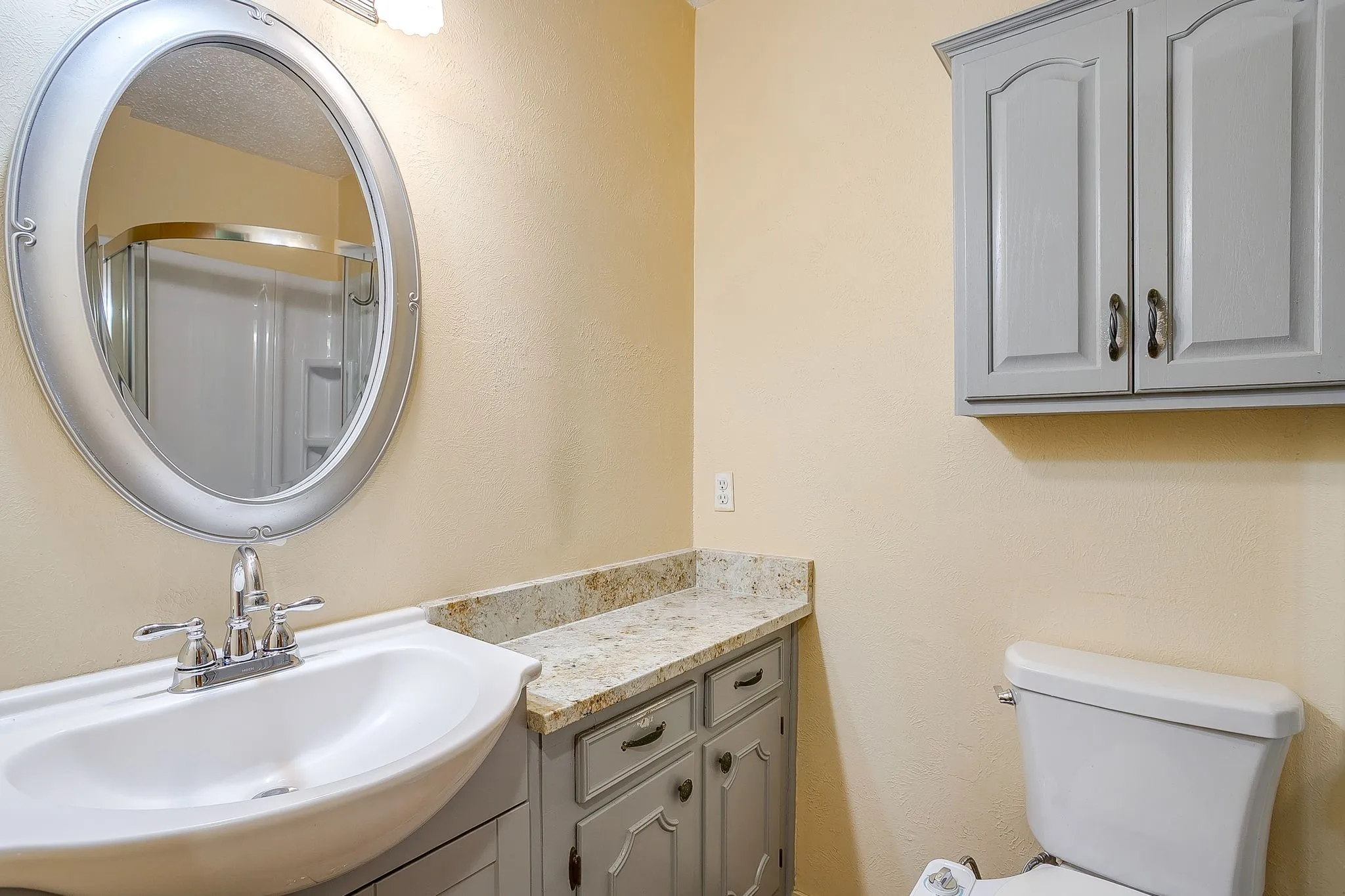 Light-toned walls, gray-painted cabinetry, a surface-mount sink, an oval vanity mirror, and a shower enclosure