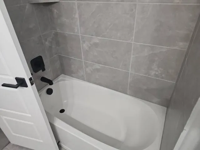 Bathtub with gray tiled surround, integrated shower niche, and matte black fixtures